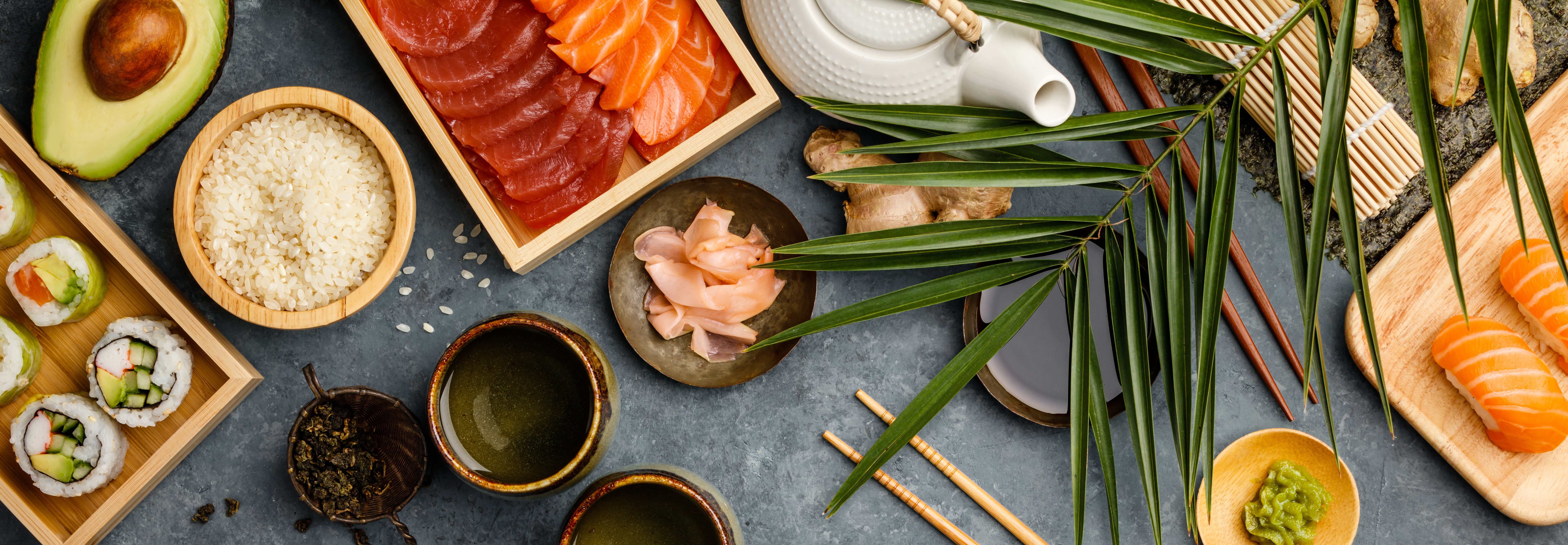 Overhead shot of ingredients for sushi, green tea and prepared maki and nigiri on dark blue background. Raw salmon and tuna pieses, rice, avocado, pickled ginger gari , raw ginger, wasabi, soy sauce, hopsticks. Asian food background. Space for text. Top view